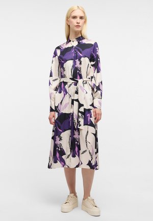 Langärmliges, kragenloses Kleid in Creme mit einem großen lila und schwarzen Blumenmuster, ausgestattet mit einem Taillenzug und einem lässigen Schnitt, kombiniert mit klobigen Sneakers.