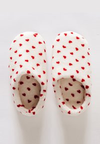 Chaussons en polaire blanche avec des motifs de cœurs rouges, présentant une texture douce et un design à dos ouvert pour faciliter l'enfilage.