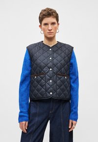 Pihe-galléros fekete mellény patentos gombokkal és barna kiemelőzsebekkel, kék kötött hosszú ujjú ing fölött viselve, sötét farmernadrággal párosítva.