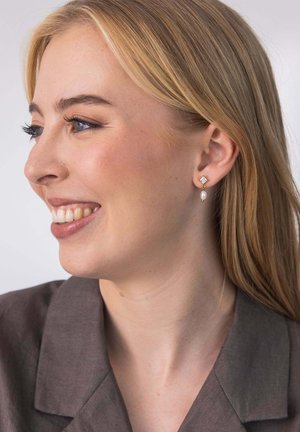 Ohrringe in Goldoptik mit einem quadratischen Stecker und einem klaren Stein sowie einer hängenden tropfenförmigen Perle. Die Haare sind glatt, blond und vom Gesicht zurückgestylt.