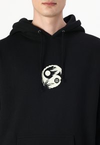 Μαύρος φούτερ με μπροστινή τσέπη, κορδόνια και κεντρικό λευκό γραφικό που απεικονίζει κυκλικά σχήματα και στοιχεία που μοιάζουν με παλάμες.