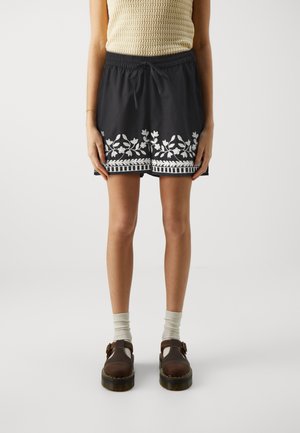 Personne portant un short noir avec une broderie florale blanche, un haut beige sans manches, des chaussettes blanches et des chaussures marron à boucle, debout sur un fond uni.