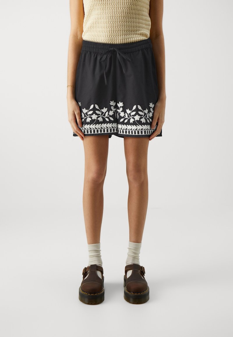 Personne portant un short noir avec une broderie florale blanche, un haut beige sans manches, des chaussettes blanches et des chaussures marron à boucle, debout sur un fond uni.