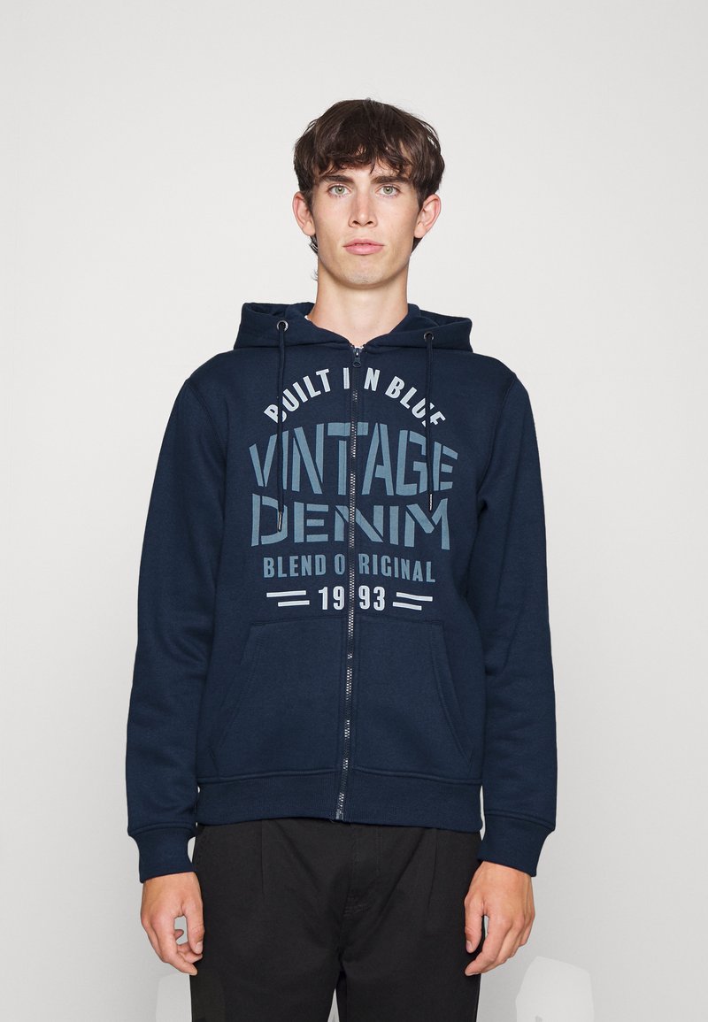 Temno modra zip-up kapuca iz mehkega materiala, ki ima sprednji grafični tisk z besedilom "Vintage Denim" in kenguru žep.