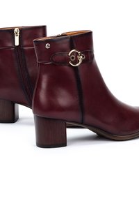 Botas de tobillo de cuero burdeos con tacón cuadrado, cremallera lateral y acento de hardware dorado en la correa. Textura suave con una costura mínima.