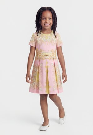 Smaidīga meitene ar pīturītēm, tērpusies rozā kleitā ar zelta izšuvumiem un valkājot baltas baleta kurpes, stāv pie baltas sienas.