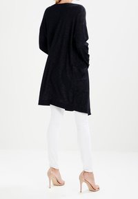 Cardigan lungo di un blu navy scuro con una vestibilità comoda, caratterizzato da una cucitura centrale sul retro e tasche laterali. Abbinato a jeans skinny bianchi e tacchi beige.