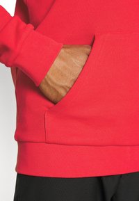 Roter Baumwollhoodie mit langen Ärmeln und gerippten Bündchen; verfügt über eine große Fronttasche. Strukturierte Oberfläche und nahtloses Design an den Rändern.