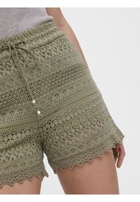 Shorts en dentelle vert olive avec taille à cordon, présentant des motifs complexes et un ourlet festonné. Fini texturé avec des accents métalliques.