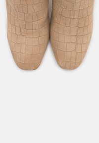 Bottes chevilles beiges avec un motif texturé imitant le crocodile, bout pointu et matière en daim doux. Semelles plates sans éléments métalliques visibles ni accents.