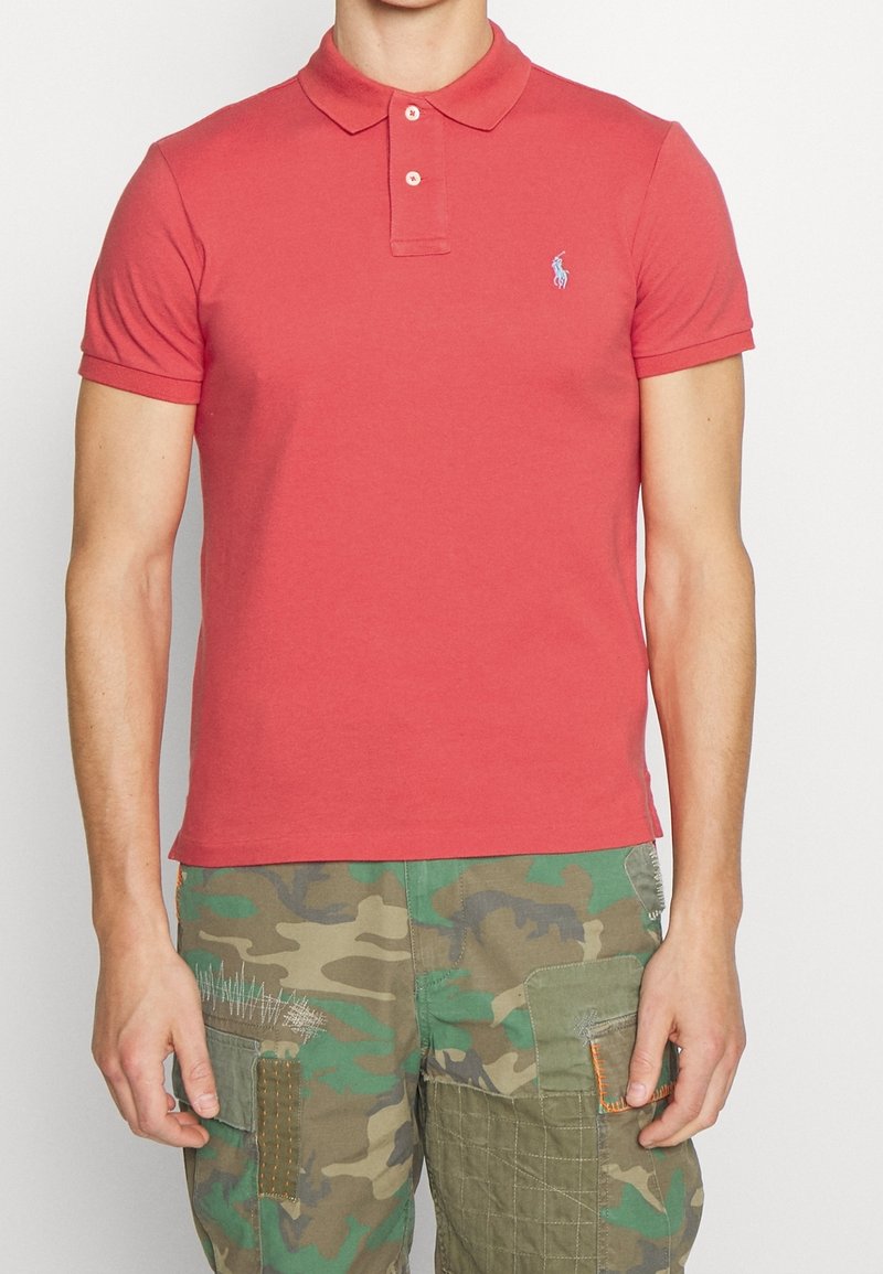 Homme portant un polo rouge à manches courtes avec un petit logo sur la poitrine gauche et un short cargo camouflage avec des détails de patchs.