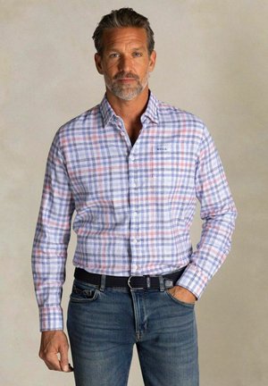 Man draagt een blauw-wit-roze geruit overhemd met knopen, ingestopt in een blauwe spijkerbroek met een zwarte riem, hand in zak, neutrale achtergrond.