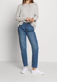 Světlé šedé pletené tričko s dlouhým rukávem spárované se středně modrými džíny s vysokým pasem a bílými teniskami. Džíny mají rovný střih a minimální detaily.