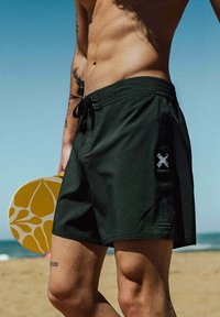 Maillot de bain noir à taille à cordon, avec un patch logo latéral et une texture lisse, tenu à côté d'une pagaie jaune aux motifs floraux.