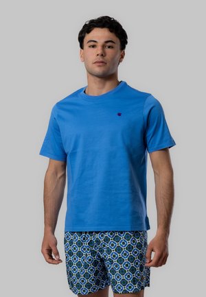 Giovane uomo in piedi che indossa una maglietta blu semplice e pantaloncini blu con fantasia su uno sfondo neutro.