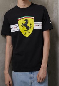 Svart bomull t-shirt med en gul Ferrari-sköldlogga, horisontella vita ränder med röda accenter över bröstet, design med rund hals.