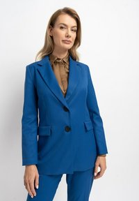 Blaues Sakko mit zwei Knöpfen, mit Revers und Fronttaschen. Kombiniert mit einem braunen Hemd mit Kragen. Glatte Textur, taillierter Schnitt.