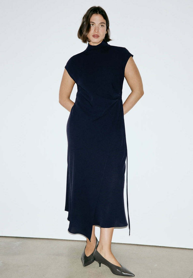 Femme debout contre un mur blanc, portant une robe midi sans manches bleu marine et des chaussures noires à bouts pointus et talons bas.