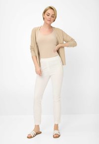 Cardigan beige léger sur un débardeur beige ajusté. Pantalons crème fuselés avec poches latérales. Sandales blanches à glissière. Palette de couleurs neutres.