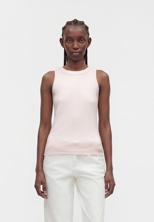 Femme aux cheveux tressés portant un haut sans manches rose clair et un pantalon blanc, debout devant un fond clair uni.