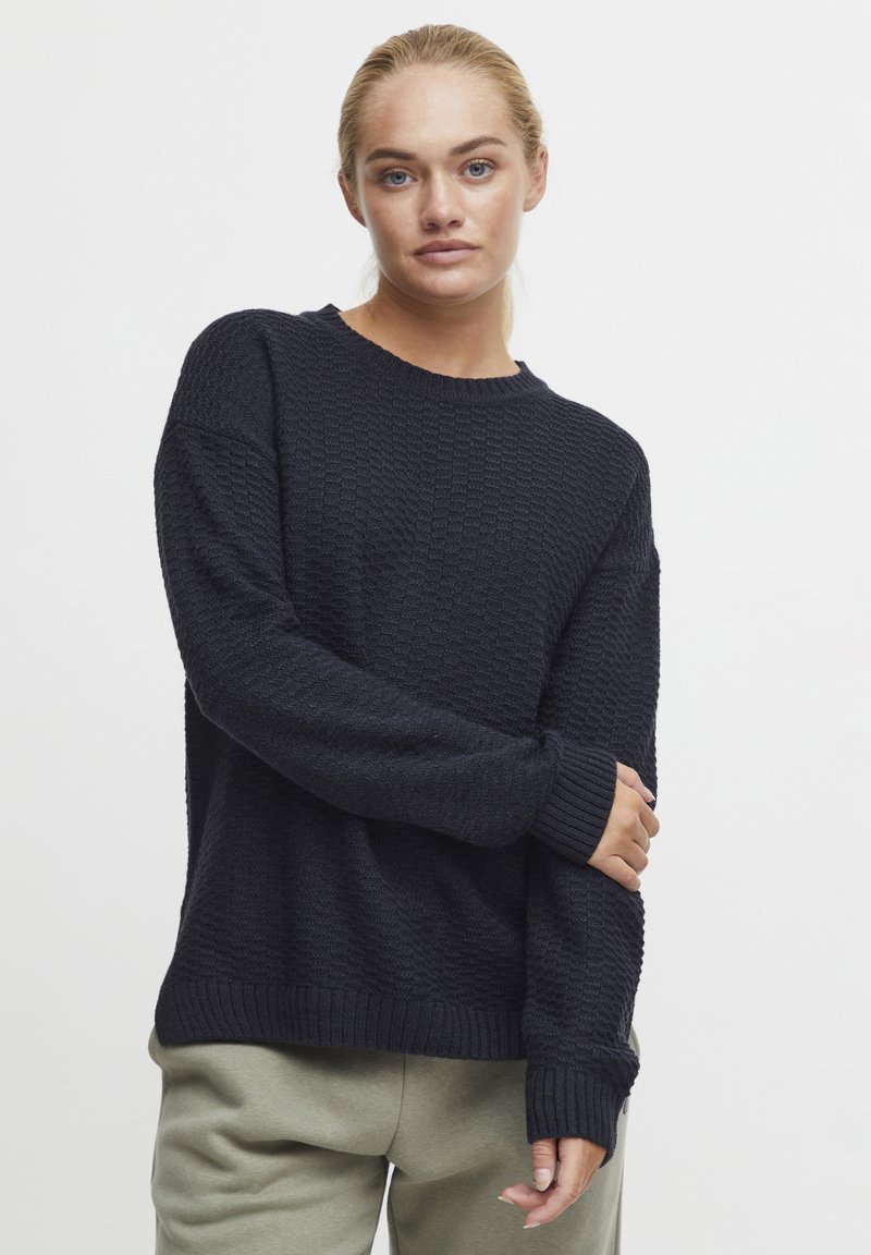Pull en maille bleu marine avec un col rond et des manches longues, comportant des poignets et un ourlet côtelés. Associé à un pantalon vert clair.