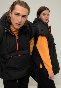 Gilet smanicati neri con accenti arancioni, chiusura con zip e tasche frontali, indossati sopra felpe arancioni e pantaloni neri. Tessuto liscio, vestibilità comoda.