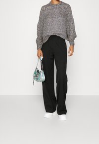 Personne portant un chemisier à manches longues à fleurs, un pantalon large noir, des chaussures blanches, et tenant un petit sac à main irisé par la poignée.