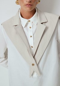 Beige layered collar jacket over a white shirt, featuring gold buttons and a smooth, tailored design. The fabric appears crisp and modern.