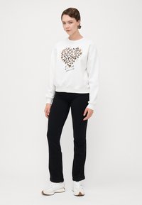 Witte sweatshirt met een hartontwerp in een luipaardprint, gecombineerd met zwarte broek en witte sneakers. Model staat met een neutrale uitdrukking.