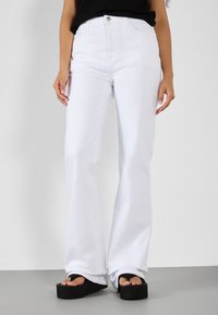 Jeans acampanados blancos de mezclilla, con una cintura alta y diseño de cinco bolsillos. Combinados con sandalias de plataforma negras y gruesas.
