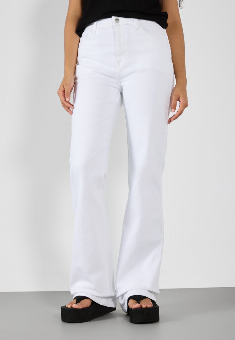 Jeans acampanados blancos de mezclilla, con una cintura alta y diseño de cinco bolsillos. Combinados con sandalias de plataforma negras y gruesas.