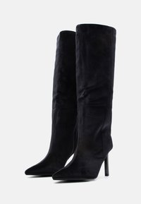 Sorte knelange støvletter med stilettheler, laget av mykt, fløyelsaktig materiale med spiss tå og slank silhuett. Har en glidelås på siden for lukking.