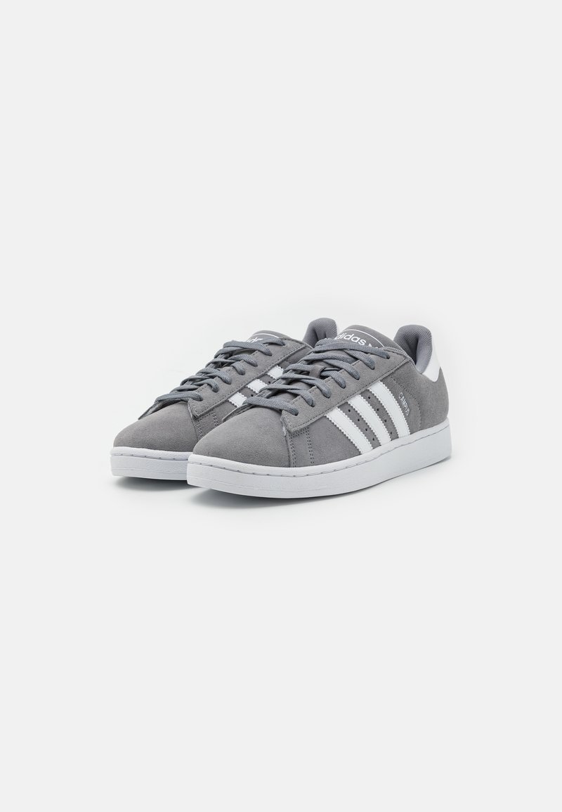 Graue Wildleder-Sneaker mit weißen Streifen, runder Spitze, flacher weißer Sohle und grauen Schnürsenkeln. Minimalistisches Design mit strukturierten Materialakzenten.