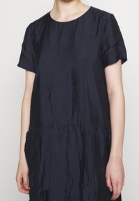 Frau in einem kurzärmeligen, dunkelblauen, locker sitzenden Kleid mit einem gerafften Bund, steht vor einem schlichten hellen Hintergrund.
