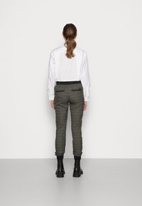 Kvinna med brun hästsvans klädd i vit långärmad skjorta, grårutiga byxor och svarta ankelstövlar står vänd mot en vit vägg.