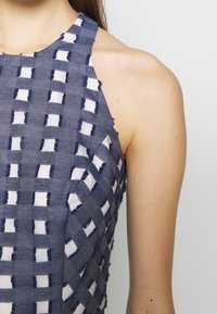 Blå och vit mönstrad tygblus med vävt design och fransade kanter, halterneck och strukturerad yta.