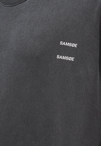 Σκούρο γκρι ύφασμα με δύο λευκά λογότυπα "SAMSØE" τυπωμένα διαγώνια στην άνω περιοχή του στήθους ενός ενδύματος.