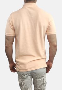 Camiseta polo de algodón en melocotón con mangas cortas, cuello y dobladillo recto, combinada con pantalones cargo en un color beige claro. La vista trasera muestra una textura suave.