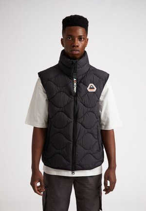 Sort quiltet vest med høj krave, lynlåslukning og logo-patch. Båret over en lys t-shirt og parret med mørke, strukturerede bukser.