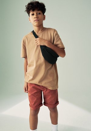 Garçon aux cheveux bouclés portant un t-shirt beige, un short rouge retroussé, des chaussettes blanches et un sac bandoulière noir, debout devant un fond uni.