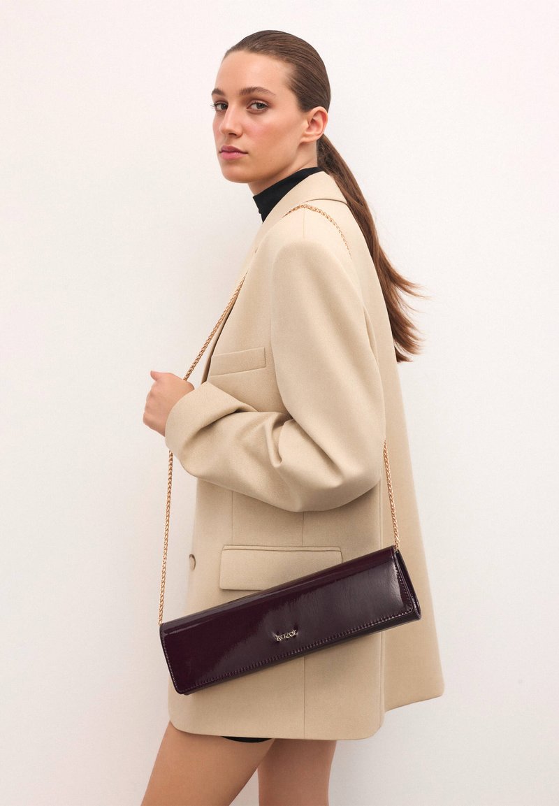 Fialová lakovaná spojka so zlatým reťazovým popruhom, obdĺžnikový tvar, hladká textura a malé logo na prednej strane.