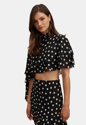 Woman with long brown hair wears black sheer polka dot crop top with layered ruffles and matching high-waisted skirt, stands against plain background.