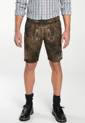 Shorts en cuir marron avec broderies décoratives, finition texturée et accents de boutons. Portés avec une chemise à carreaux et des chaussures noires.