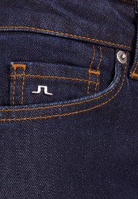 Jeans de mezclilla azul oscuro con costuras naranjas, que presentan un pequeño logo plateado sobre el bolsillo delantero y un botón metálico en la cinturilla.