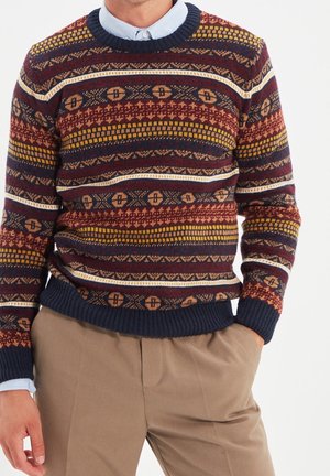 Mann trägt einen marineblauen, bordeauxroten, senffarbenen und beigen gemusterten Pullover über einem hellblauen Hemd mit Kragen und beigen Hosen, Hände entspannt an den Seiten.