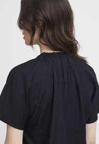 Haut noir à manches courtes avec un tissu texturé, un col froncé et une coupe ample. Coutures visibles et détail drapé au dos.