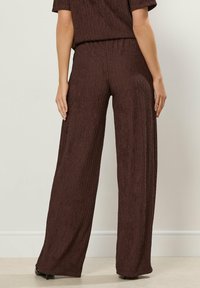 Femme portant un pantalon large texturé marron foncé et un haut assorti, se tenant contre un mur uni couleur ivoire.