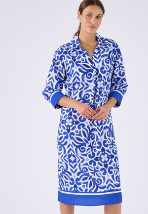 Mujer vestida con un vestido azul y blanco de manga larga y cuello, patrón hasta la rodilla, de pie con las manos entrelazadas al frente.