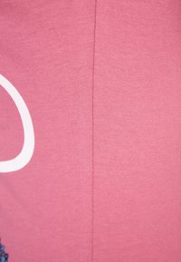 Rosa tyg med en slät textur, som har ett vitt designelement och fina sömnadsdetaljer längs en söm.