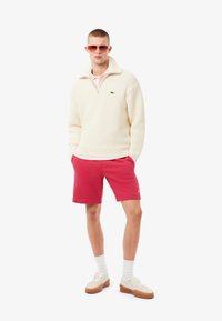 Suéter de punto crema con collar alto, combinado con pantalones cortos rosas brillantes. Zapatillas beige, calcetines blancos y gafas de sol con tinte naranja completan el conjunto.